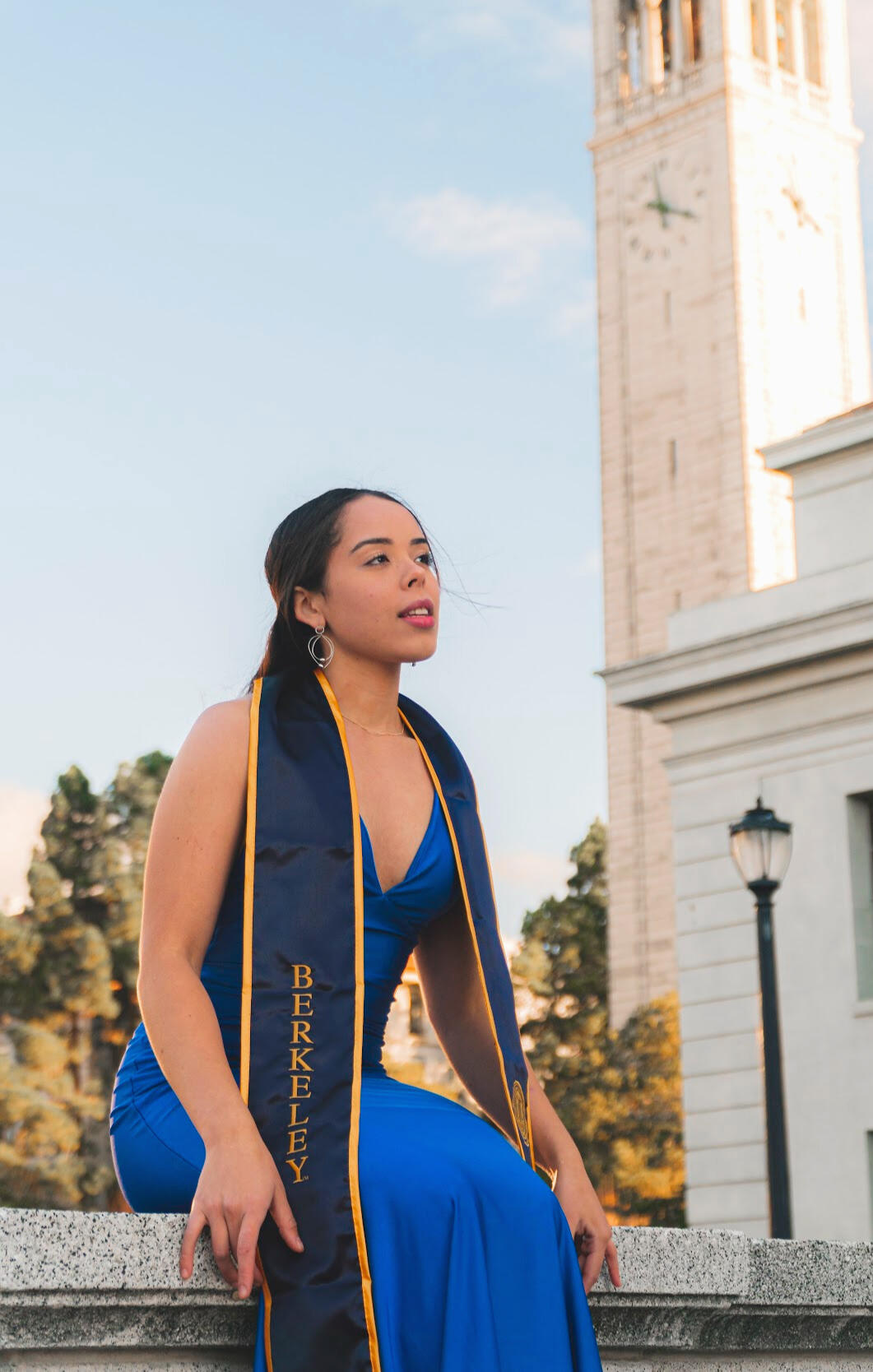 The Graduation An excited young girl in a vibrant blue dress, adorned with UC Berkeley stoles, sitting on a bench with the iconic UC Berkeley clock tower in the background