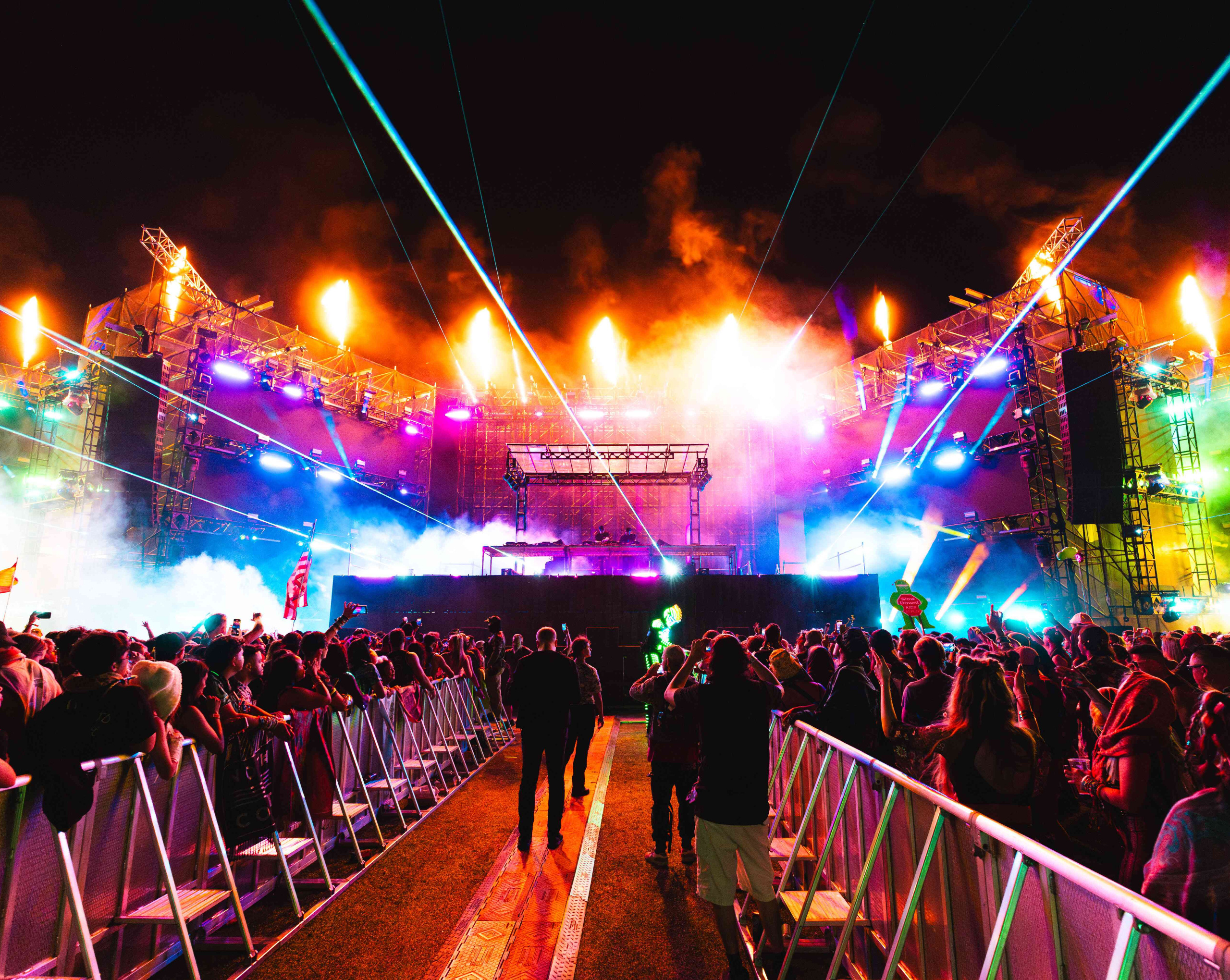 Slander performing on the main stage with a massive crowd, surrounded by multicolored lights, lasers, and a dramatic flame show.